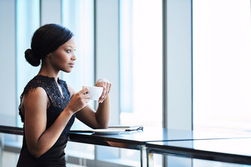 Elegant African American woman thoughtfully looking out the large window while holding her cup of tea in her hands, resting one elbow on the counter maintaining regal posture - Powered by Adobe