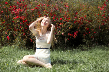 Young blonde woman in lacy white dress enjoying aroma blooming garden in the air. Girl sitting on fresh grass