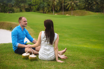 Young beautiful couple with coconuts holding their hands at golf course in summer. man wear the blue shirt and the girl in a white dress. Concept of honeymoon