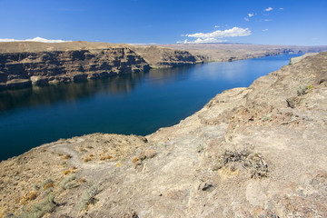 Washington state landscape with Columbia river and canyon. Geological formation with rocky shores with sparse vegetation and bright water reflecting the blue sky.