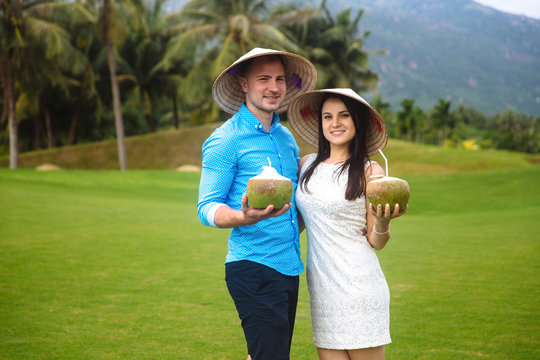 Young beautiful couple in love holding coconut in hands and walking at golf course in summer. man wear the blue shirt and the girl in a white dress and vietnamese hats. Concept of honeymoon