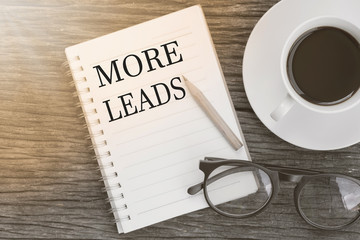 Concept MORE LEADS message on notebook with glasses, pencil and coffee cup on wooden table.