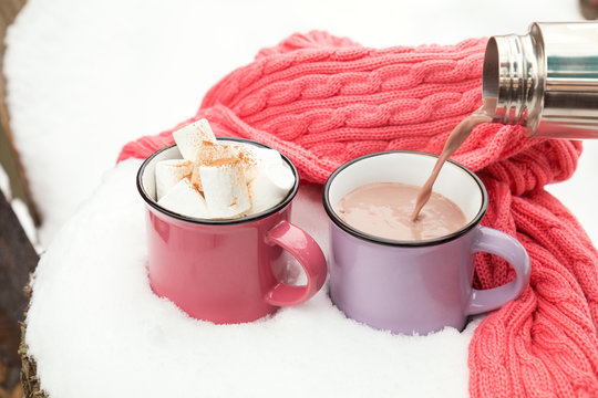 Pouring hot chocolate in the cup. Hot chocolate with marshmallow in pink and violet two cups wrapped in a cozy winter pink scarf on the snow-covered table in the garden. Coloring and processing photo