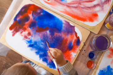 Close-up of watercolor wet-on-wet paints with little boy's head and hand holding paintbrush and drawing in Waldorf kindergarten