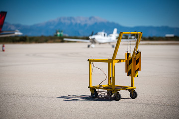 A yellow aircraft chocks and fire extinguisher carrier on a small airport. Aircraft chocks are hanging on the carriers handle. Small aircraft and scenic view on the background.
