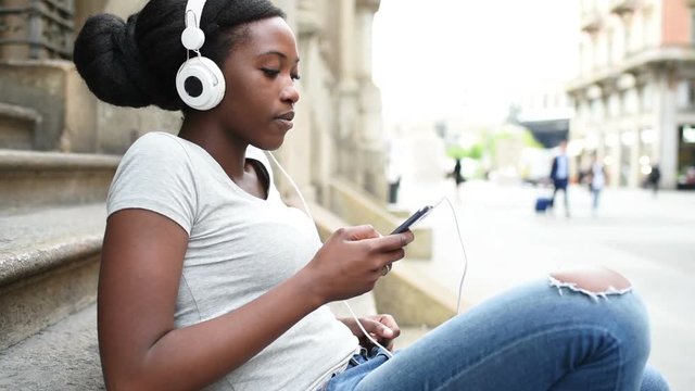 Young beautiful black woman outdoor listening music with head phones and smart phone hand hold, smiling - relaxing, happiness, technology concept