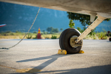 A rear landing gear and wheel chocks of a small aircraft on the ground with blurry nature and mountain in the background. - obrazy, fototapety, plakaty