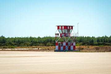 A small white and red air traffic control tower next to the empty airport runway. Green fields and blue sky in the background.