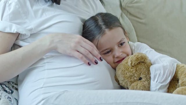 Tracking shot of little girl laying on belly of her pregnant mother and holding teddy bear