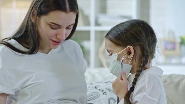 Tilt down of little girl playing with pregnant mother as doctor and patient. She wearing medical face mask and using stethoscope to listen to heartbeat of baby in belly of woman