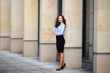 Brunette smiling woman standing near the wall of building, looking forward into camera