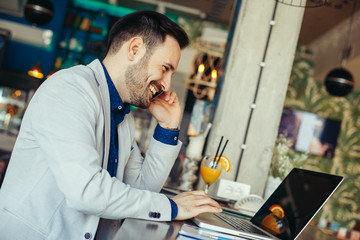 Fototapeta premium Young businessman talking on the phone and looking at laptop on table in cafe