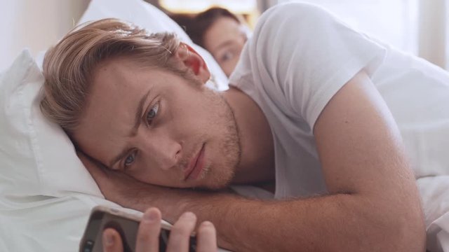 Handsome bearded young male wearing white shirt lying upset in bed in the morning looking at mobile phone with his girlfriend next to him in slowmotion