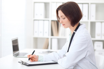 Young brunette female doctor sitting at the table and working at hospital office.  Health care, insurance and help concept. Physician ready to examine patient