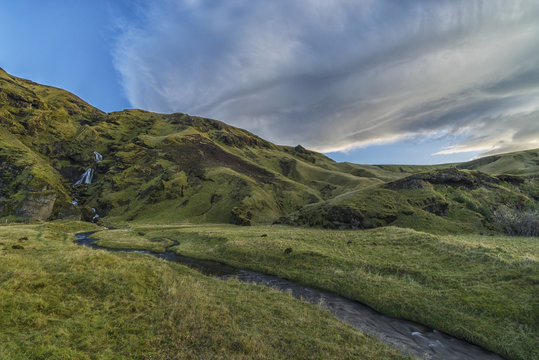 Lenticular clouds form over a waterfall and stream running towards the ocean near Vik; Iceland