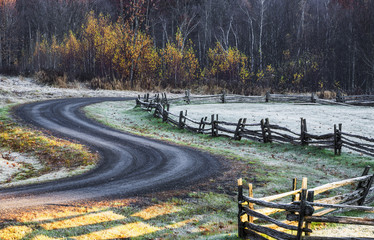 Wooden rail fence in a frost covered grass field with trees in autumn colours and a winding road; Iron Hill, Quebec, Canada