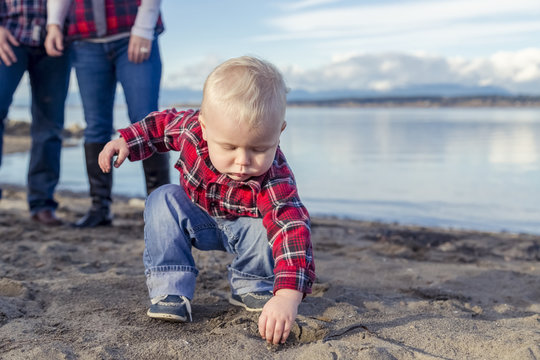 A young toddler gets his hands dirty playing with sand on the beach while his parents look on from the background; Surrey, British Columbia, Canada
