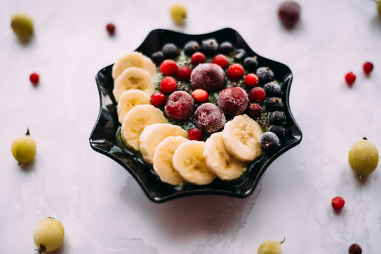 Green Smoothie, Decorated With Slices Of Banana, Cherries, Currants, Viburnum, Sesame Seeds And Chia Seeds. Smoothies In A Black Bowl On A White Background. The Berries Lie Around