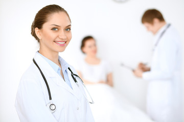 Fototapeta premium Cheerful smiling female doctor on the background with physician and his patient in the bed. High level and quality of medical service concept.