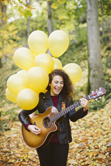 Beautiful young woman playing guitar on forest with yellow balloons. Girl wearing black jacket, fashion lifestyle.