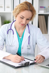 Happy blonde female doctor sitting at the table and filling up medical history form. Medicine, healthcare and help concept. Physician ready to examine patient