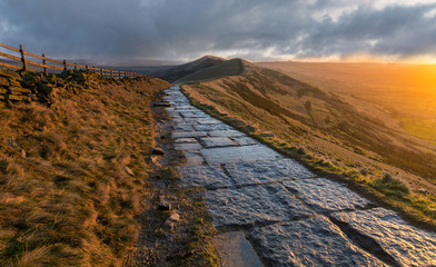 Stone footpath leading a long Mam Tor mountain ridge with moody storm clouds and golden light.