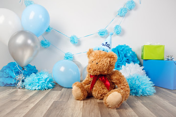 Festive background decoration for first year birthday celebration with little bear toy sitting on floor in studio, balloons, paper flowers and presents gift boxes