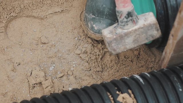 Slowmotion sledgehammer hitting piece of wood stuck between two black plastic pipe on sand. Close up