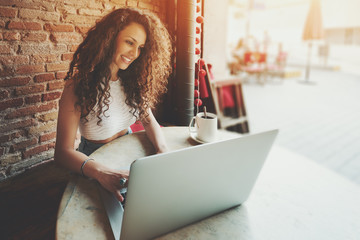 Smiling curly female student working on net-book with blank copy space screen for your text message or promotional content, successful freelancer woman is using laptop computer while sitting in cafe