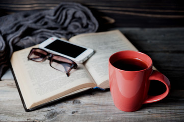 grey cozy knitted scarf with cup of coffee or tea, phone, glasses and open book on a wooden table.
