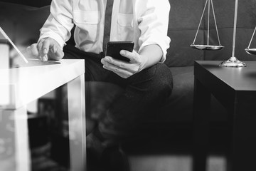 Justice and Law context.Male lawyer hand sitting on sofa and working with smart phone,digital tablet computer docking keyboard with gavel and document on living table at home,black and white