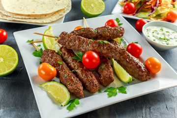 Homemade Kofta kebabs on skewers with pita, lime, vegetables, sweet chili and yogurt sauce on white plate.