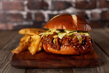 Pulled pork burger on pretzel bun with potato wedges on a wood server with brick background