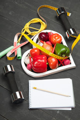 Sport and diet. Healthy lifestyle. Vegetables, dumbbells  notebook. Peppers, tomatoes, garlic, onion  radish in a heart on  black background