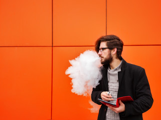 Young hipster man with a beard and glasses smokes an electronic cigarette. He holds a tablet pc. Youth fashion. Delicious pairs. A cloud of steam.