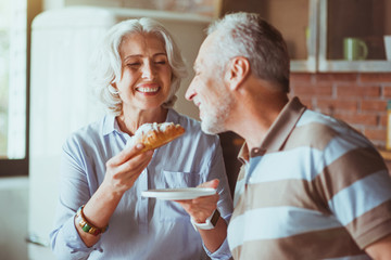 Cheerful aged woman giving croissant to her husband