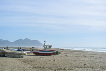 Fototapeta premium Cloudy day at the lonely beach with colored boats on the shore not far from mountains.