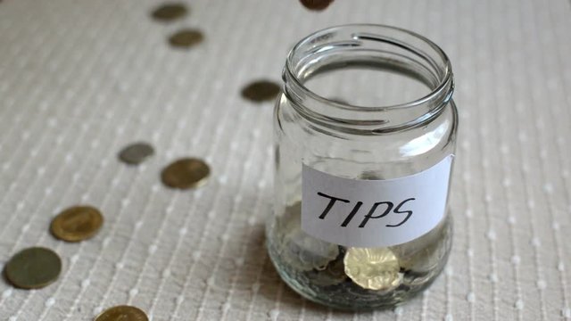 Coins filling and overflowing a clear medium sized glass jar labeled for tips.