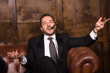 Rich businessman with cigar sitting on sofa and looking at camera. Happy man spending free time in...