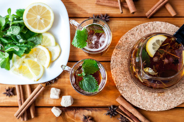 Clear glass and teapot of hot black herbal tea with lemon, mint and Melissa leaves on light rustic wooden table. Summer, Autumn, winter drink. Cinnamon and cane sugar for decoration. Top view.