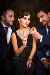 Focus on pretty lady with red lips sitting with glass of champagne. Two handsome rich executive businessmen have rivalry or competition for elegant lady in restaurant atmosphere.