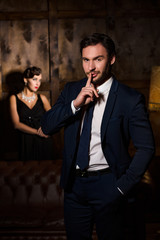 Wealth, luxury concepts. Handsome man asking for silence and keeping hand in pocket. Elegant gorgeous lady sitting on background.