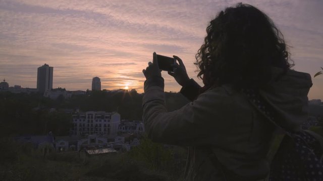 Inspired female student filming cityscape with amazing sunset on smartphone