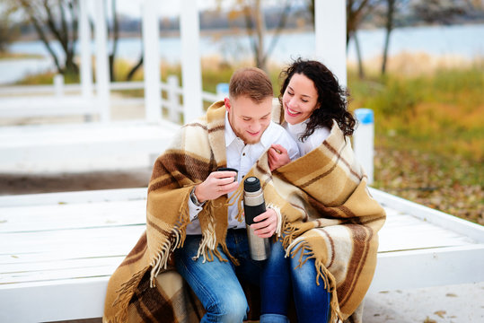 Young couple embracing and covered with a warm blanket sitting on autumn beach and drink coffee from a thermos