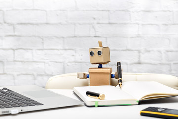 robot is sitting at a table, holding a pen and looking at a laptop