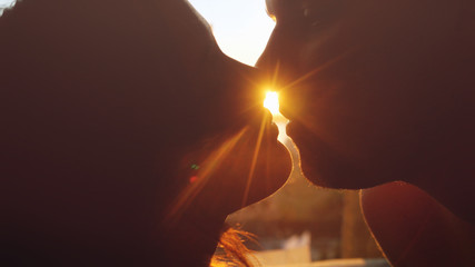Romantic young beautiful couple in love looking each other and kissing on a sunset with sun shining bright behind them on a horizon.