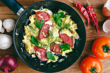 Fresh cooked scrambled eggs in pan with sausage, herbs. Tomato, chilli pepper, onion, mushroom on wooden board, top view