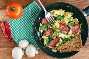 Fresh cooked scrambled eggs in pan with sausage and herbs. Tomato, chilli pepper, mushrooms on wooden board, top view.