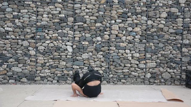 Young male trendy hip-hop dancer outside, doing break-dance in city setting in front of stone wall