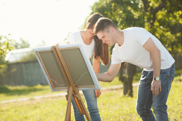 Young couple drawing at easel by colorful variegated paints. Pretty young woman and handsome guy having fun with paints outdoors.